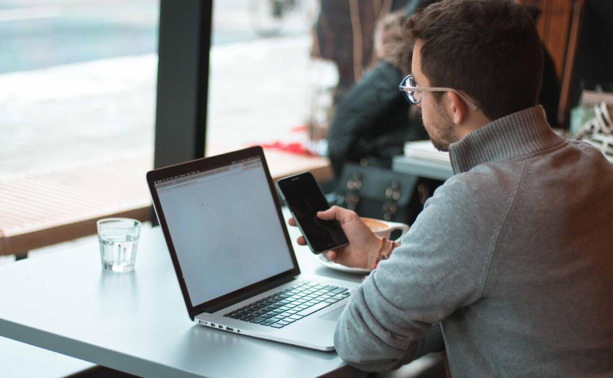 Man in a casual sweater working on a laptop while checking his smartphone in a cafe.