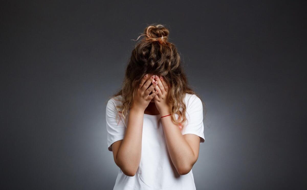 Young beautiful girl hiding face with hands over grey wall.